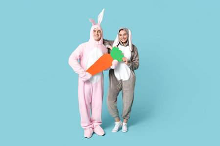 Beautiful young couple in bunny costume with paper carrot on blue background. Easter celebration