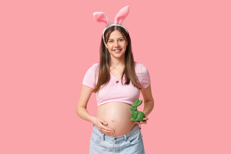 Beautiful young pregnant woman in bunny ears with toy rabbit on pink background. Easter celebrationの写真素材