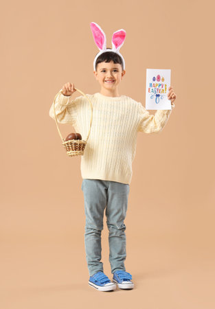 Cute little boy in bunny ears headband holding basket with chocolate eggs and festive postcard for...