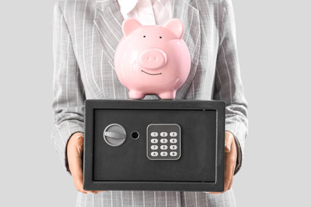 Beautiful young Asian woman with safe box and piggy bank on gray background, closeup