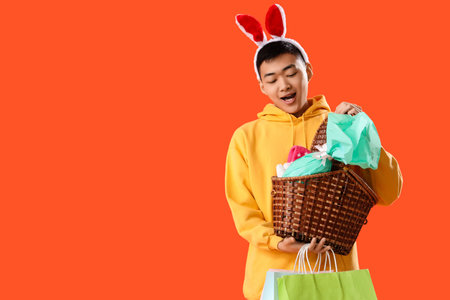 Young Asian man in bunny ears with basket and shopping bags on orange background. Easter Saleの写真素材