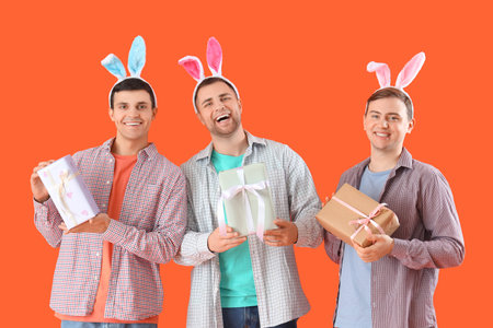 Happy young men in bunny ears with gift boxes on orange background. Easter celebration