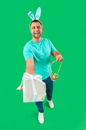 Happy young man in bunny ears with gift box and wicker basket on green background. Easter...