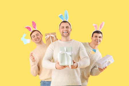 Happy young men in bunny ears with gift boxes and paper decorations on yellow background. Easter celebrationの写真素材