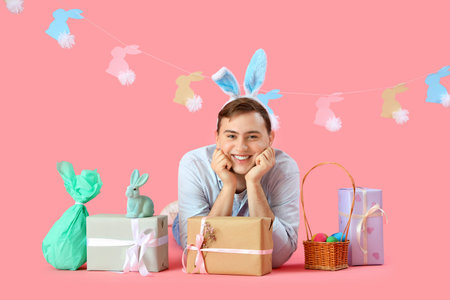 Happy young man in bunny ears with gift boxes, wicker basket and rabbits garland on pink background. Easter celebrationの写真素材