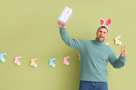 Happy young man in bunny ears with gift box and rabbits garland showing victory gesture on green...