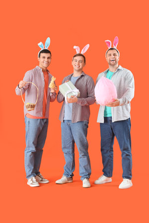 Happy young men in bunny ears with gifts and wicker basket on orange background. Easter celebration