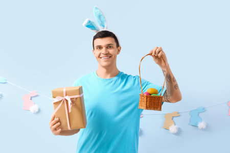 Happy young man in bunny ears with gift box, wicker basket and rabbits garland on blue background....