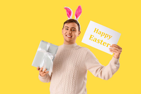 Happy young man in bunny ears with gift box and greeting card on yellow background. Easter...