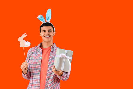Happy young man in bunny ears with gift box and paper rabbit on orange background. Easter celebrationの写真素材