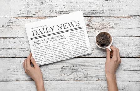 Female hands with newspaper and cup of coffee on white wooden background. Top view