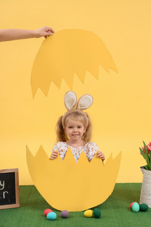 Cute little girl in paper egg shell with Easter eggs on yellow background