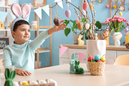 Cute little boy in bunny ears decorating willow branches with Easter eggs at home