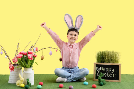 Cute little boy in bunny ears with Easter eggs, toy rabbit and decorations sitting on yellow...