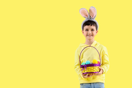 Cute little boy in bunny ears holding basket with Easter eggs on yellow background