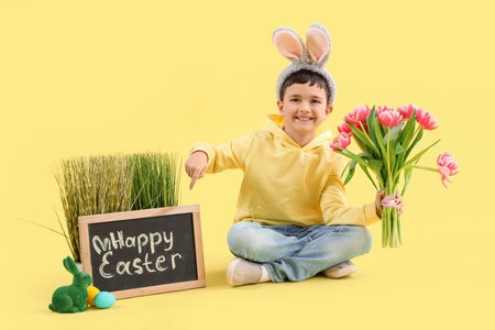 Cute little boy in bunny ears holding tulip flowers and pointing at chalkboard with text HAPPY...