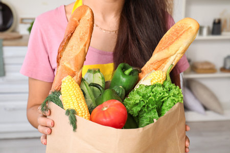 Beautiful young Asian woman with bag full of fresh products in kitchenの写真素材