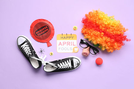 Shoes with tied together laces, clown wig and festive postcard for April Fools Day on lilac...