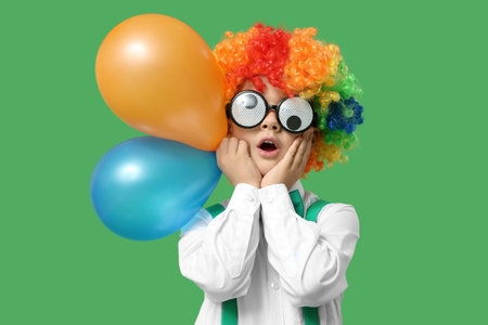 Surprised little boy in funny glasses and clown wig with balloons on green background. April Fools Day celebrationの写真素材