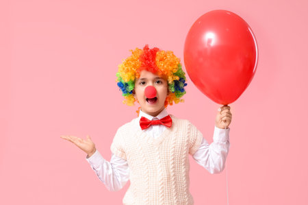 Surprised little boy in clown wig and nose holding balloon on pink background. April Fools Day celebrationの写真素材