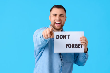 Young man holding paper with text DON'T FORGET and pointing at viewer on blue backgroundの写真素材