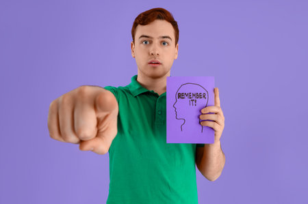 Young man holding paper with text REMEMBER IT and pointing at viewer on lilac background