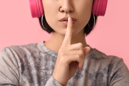 Stylish young woman in headphones with nose piercing showing silence gesture on pink background