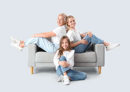 Little girl with her mom and grandmother sitting on sofa against light background