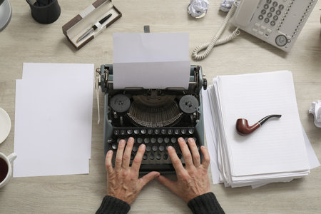 Mature man with paper sheets and smoking pipe typing on vintage typewriter at beige wooden table