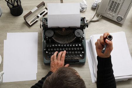 Mature man with paper sheets and smoking pipe typing on vintage typewriter at beige wooden table