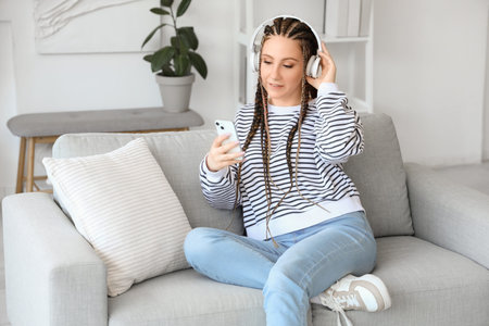 Young woman with dreadlocks in headphones using mobile phone at home
