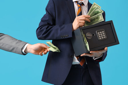 Young man with safe box and hand holding money on blue background, closeup