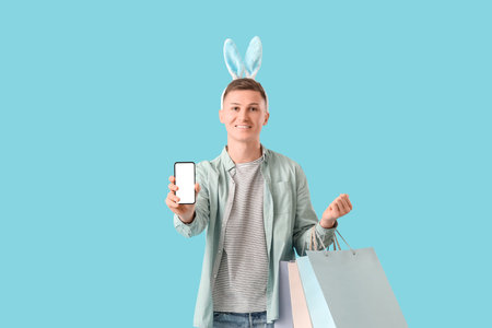 Young man in bunny ears with mobile phone and shopping bags on blue background. Easter Sale