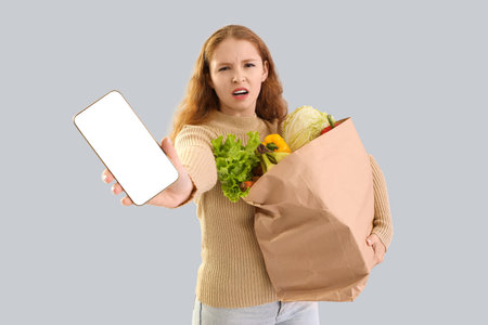 Displeased young woman with paper bag of healthy food and mobile phone on light backgroundの写真素材
