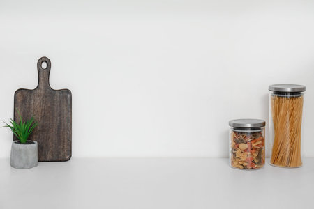 Jars with different types of pasta, wooden board and houseplant on white kitchen counter