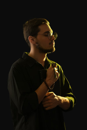 Young man with rosary beads and Bible praying on black background