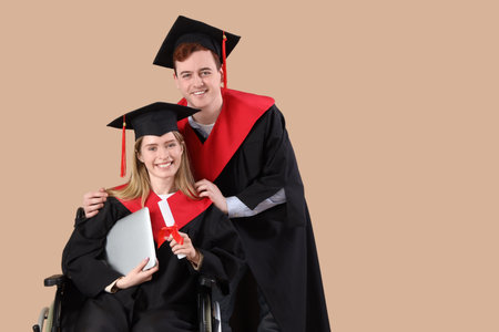 Male graduate in wheelchair with laptop and classmate on beige background