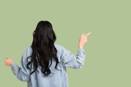 Beautiful young woman with dark wavy hair in denim jacket pointing at something on green background,...