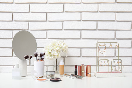 Different makeup products, mirror and beautiful flowers on dressing table near light brick wall in roomの写真素材