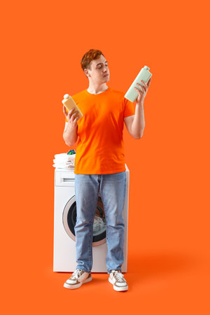 Young man with detergents near washing machine on orange backgroundの写真素材