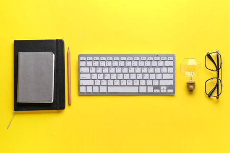 Composition with modern computer keyboard, light bulb, eyeglasses, notebooks and pencil on yellow backgroundの写真素材