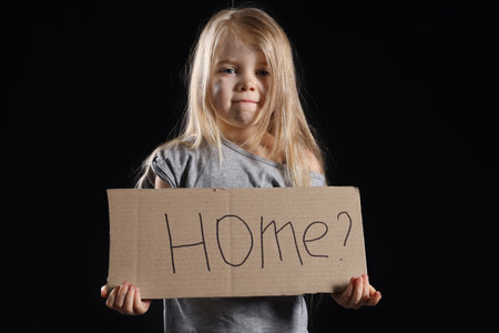 Homeless little girl holding piece of cardboard with word HOME on dark backgroundの写真素材