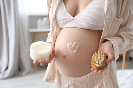Young pregnant woman with heart made of cream on her belly and massage brush in bedroom, closeupの写真素材