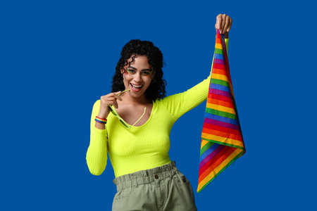 Happy African-American woman with rainbow flag and stylish sunglasses on blue background. LGBT...
