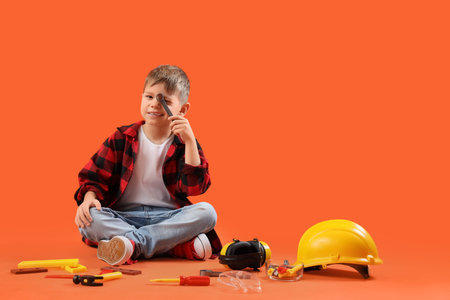 Cute little builder with toy tools sitting on orange background