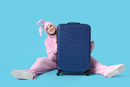 Young man in bunny costume with suitcase sitting on blue background. Easter celebrationの写真素材