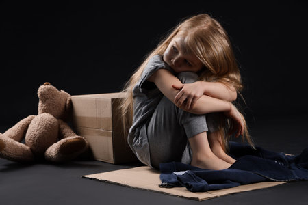 Homeless little girl sitting on piece of cardboard against dark backgroundの写真素材