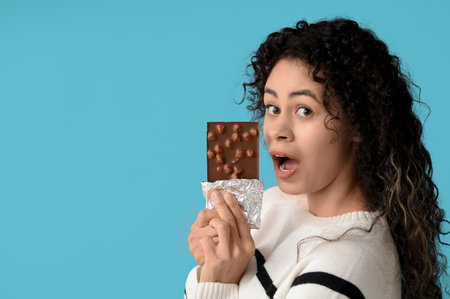 Shocked young African-American woman with sweet chocolate bar on blue backgroundの写真素材