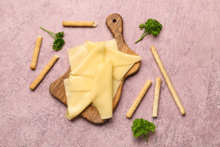 Board with tasty cheese slices, parsley and Italian Grissini on pink background