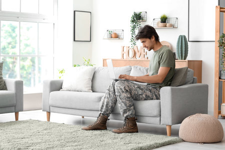 Young male soldier with laptop sitting on sofa at homeの写真素材